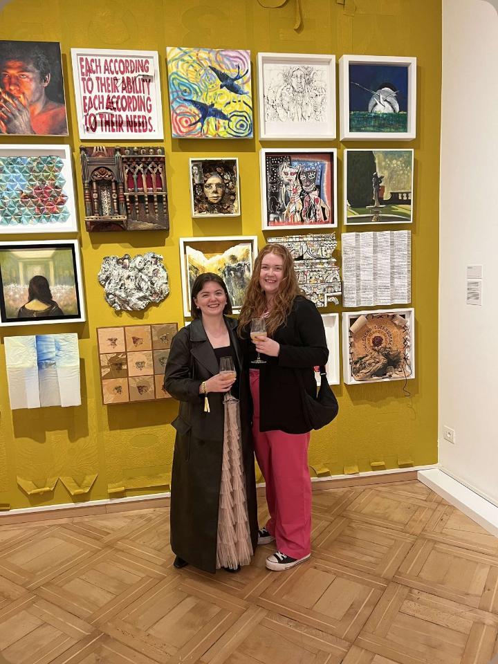 Rectangular photo - PORTRAIT - Optimised for web, Student Success Story - picture of two females next to each other with their arms around each other - both holding a glass and smiling. They are standing in front of a gallery wall.