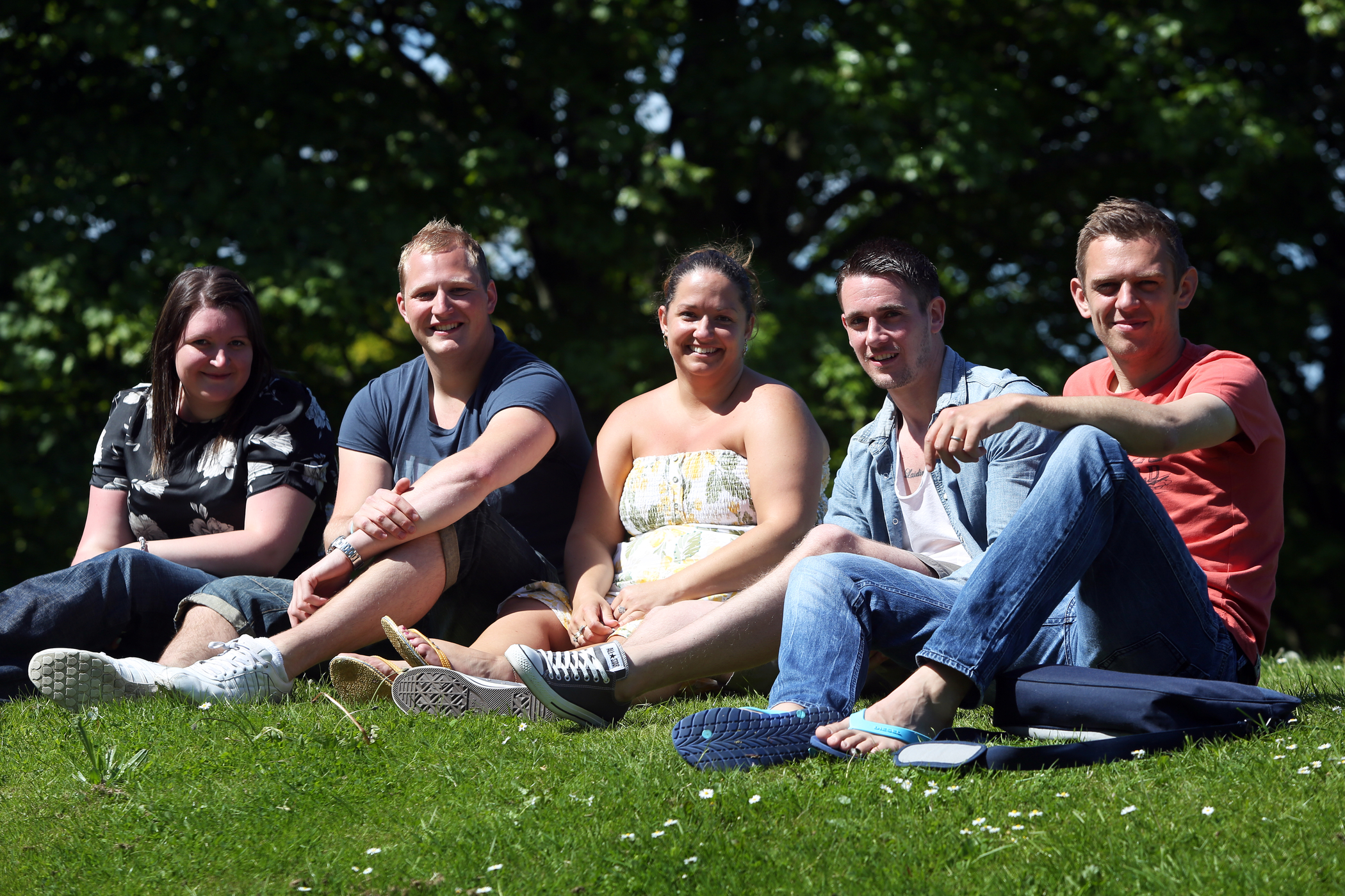 A group of people sitting on the grass enjoying the sunshine and smiling at the camera.