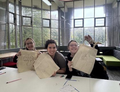 Crafternoon - Lancaster , The image shows three students holding up tote bags they have painted, they are sat in the Library at Lancaster Campus and are attending a Student Services crafternoon session