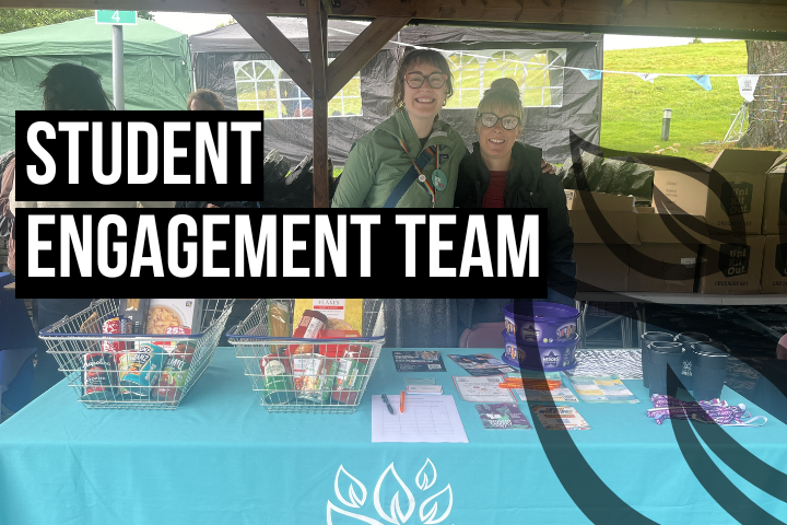 The image shows two staff members stood behind a table at Ambleside's 2025 welcome fair, with the text 