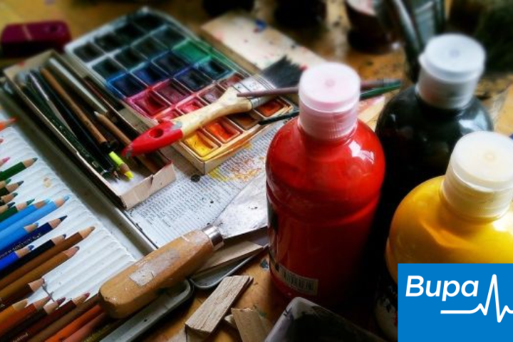 The image shows a messy desk full of art supplies like, bottles of acrylic paint, a watercolour palette and coloured pencils. The 