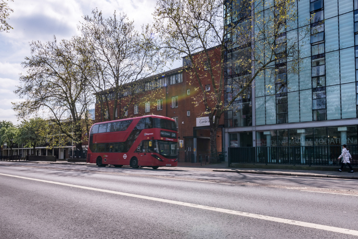 A red double-decker bus travels along a wide city street lined with modern office buildings and mature trees. The scene features a glass-fronted building alongside a brick structure displaying signage for Cumbria House. Sunlight filters through the trees, creating lens flare and casting shadows across the road. The urban streetscape combines contemporary architecture, public transport, and pedestr