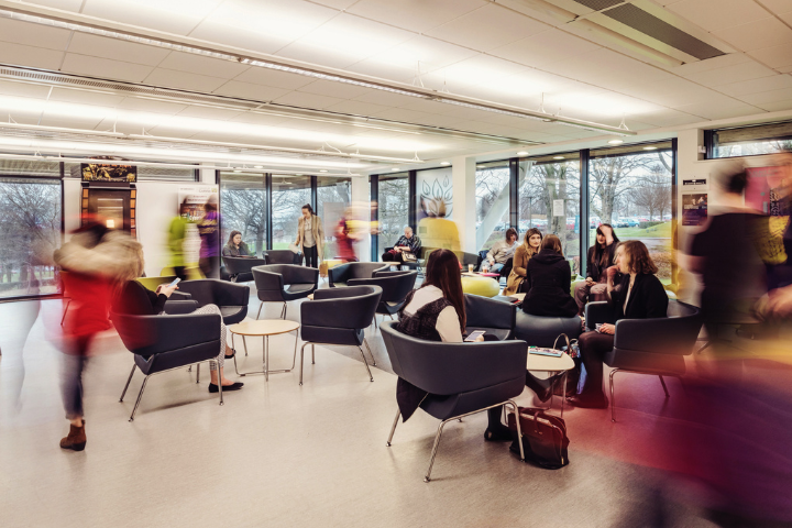 Students gather and socialise in a modern communal area at the University of Cumbria. The bright, open space features comfortable chairs, small tables, and large floor-to-ceiling windows overlooking trees and outdoor greenery. Some students are seated in conversation while others move through the lounge, creating a sense of activity and community. Natural light fills the contemporary study and soc