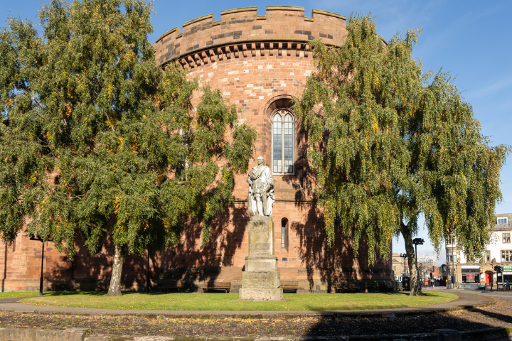 A historic red sandstone building with a circular fortified tower stands in Carlisle, Cumbria, framed by mature trees with cascading branches. In the foreground, a stone statue on a pedestal is positioned on a grassy area, while the castle-style architecture features crenellated walls and a tall arched window. Bright sunshine and a clear blue sky illuminate the scene, highlighting the textures of