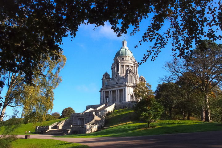 The Ashton Memorial stands atop a grassy hillside in Williamson Park, Lancaster, beneath a bright blue sky. The grand white stone monument features a large green copper dome, classical columns, and ornate architectural detailing. Curving staircases lead up through landscaped gardens and mature trees, while overhanging branches frame the scene. The striking Edwardian landmark is surrounded by well-