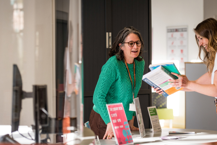 University staff member assisting a colleague at a student information desk, with brochures and university resources displayed on the counter.