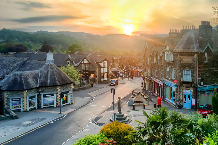 Elevated view of Ambleside town centre in the Lake District, Cumbria, at sunset. Stone-built historic buildings and shops line a quiet road leading through the town, with warm sunlight breaking over the hills in the background. The scene features traditional architecture, a small central monument, lush greenery and palm-like plants in the foreground, with rolling fells and a golden sky creating a
