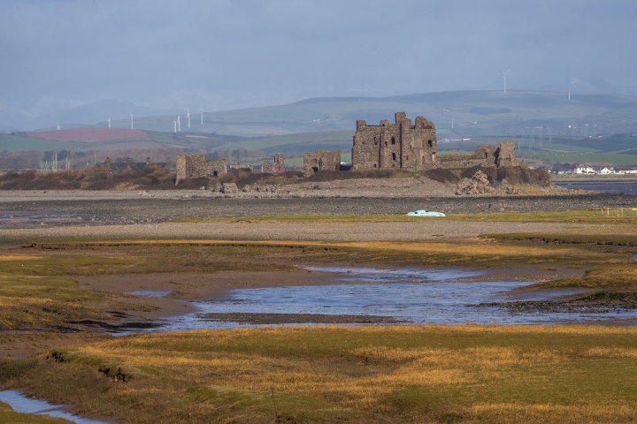 Piel Castle ruins stand on a low island off the coast of Barrow-in-Furness, Cumbria, surrounded by tidal flats and shallow water channels. The foreground features marshland and exposed shoreline at low tide, while rolling hills and several wind turbines are visible in the distance beneath a cloudy sky. The historic stone remains of the medieval fortress form the focal point of this tranquil coasta