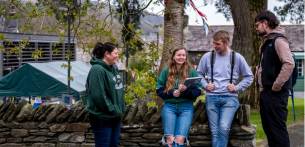 Four university students gathered outdoors beside a traditional stone wall on the University of Cumbria campus. Two students are seated on the wall holding notebooks and drinks, while two others stand nearby engaged in conversation. Trees, campus buildings, and green outdoor spaces create a welcoming and community-focused university environment.