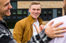 The image shows an international student smiling. In the foreground of the image another international student has their hand on another students back.