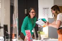The image shows a library staff member at the StEP desk at Lancaster campus, smiling and helping a student.