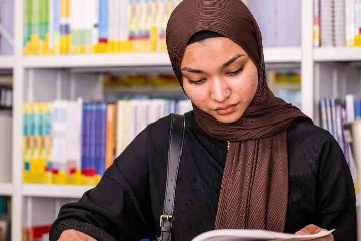 University student studying from a book in a university library surrounded by bookshelves.