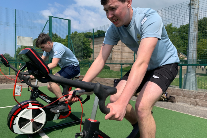 Two people using stationary exercise bikes on an outdoor sports court surrounded by fencing on a sunny day. One person is in the foreground riding a bike with red and black accents, while another person cycles on a similar bike in the background. Green artificial turf, trees, and blue sky with light clouds are visible behind the training area.